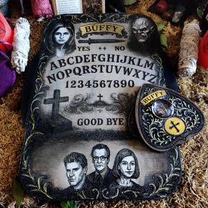 May include: A decorative Ouija board with the word "Buffy" at the top, featuring portraits of characters and the alphabet. The planchette is heart-shaped with a cross design. The board is black and cream-colored.