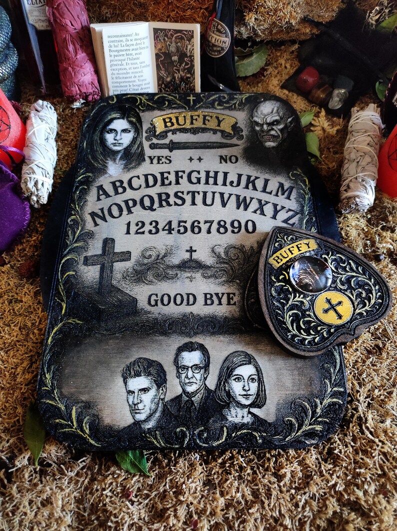 May include: A decorative Ouija board with the word "Buffy" at the top, featuring portraits of characters and the alphabet. The planchette is heart-shaped with a cross design. The board is black and cream-colored.