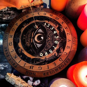 May include: A wooden Ouija board with a black and white eye design in the center. The board has a circular shape and is surrounded by candles. The board has letters, numbers, and the words "Oui" and "Peut-&ecirc;tre" printed on it.