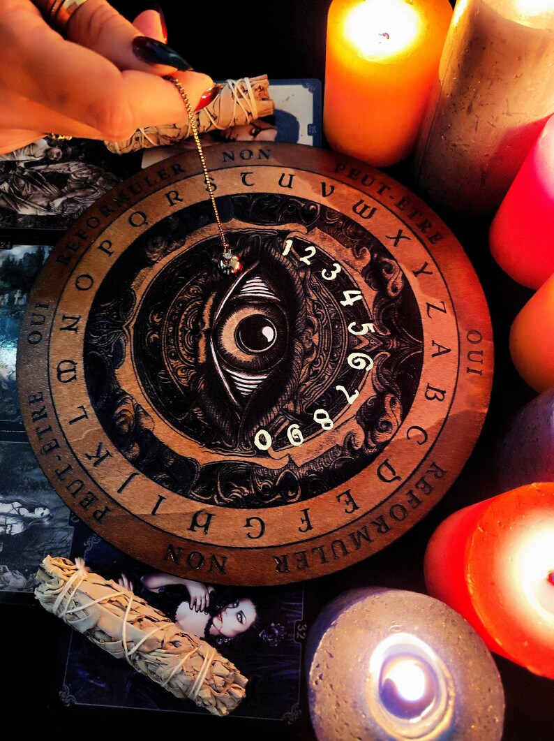 May include: A wooden Ouija board with a black and white eye design in the center. The board has a circular shape and is surrounded by candles. The board has letters, numbers, and the words "Oui" and "Peut-&ecirc;tre" printed on it.