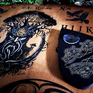 May include: A wooden Ouija board with a black and white illustration of a tree with a face. The board has the letters of the alphabet, numbers, and the words "yes", "no", "goodbye", and "au revoir". A black planchette with gold accents and a blue glass orb is on top of the board.