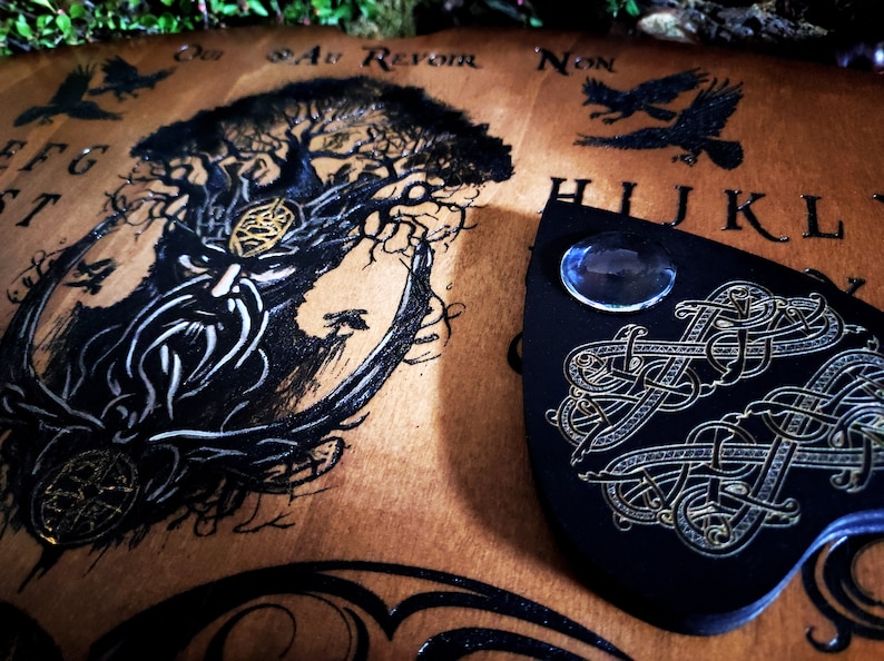May include: A wooden Ouija board with a black and white illustration of a tree with a face. The board has the letters of the alphabet, numbers, and the words "yes", "no", "goodbye", and "au revoir". A black planchette with gold accents and a blue glass orb is on top of the board.