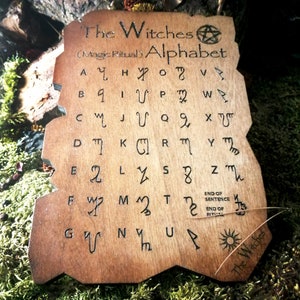 Puede incluir: Una placa de madera con una mancha marr&oacute;n y un dise&ntilde;o quemado del alfabeto de las brujas. El texto dice "The Witches (Magic Ritual) Alphabet" e incluye un s&iacute;mbolo de pent&aacute;culo. El alfabeto est&aacute; escrito en una fuente estilizada con cada letra correspondiente a un s&iacute;mbolo.