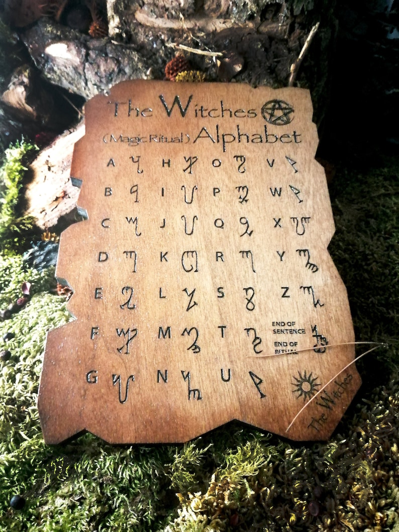 Puede incluir: Una placa de madera con una mancha marr&oacute;n y un dise&ntilde;o quemado del alfabeto de las brujas. El texto dice "The Witches (Magic Ritual) Alphabet" e incluye un s&iacute;mbolo de pent&aacute;culo. El alfabeto est&aacute; escrito en una fuente estilizada con cada letra correspondiente a un s&iacute;mbolo.