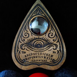 May include: A wooden Ouija board with engraved letters, numbers, and decorative elements. The board features a planchette and the words "Yes" and "No". The board is brown with black engraved details.