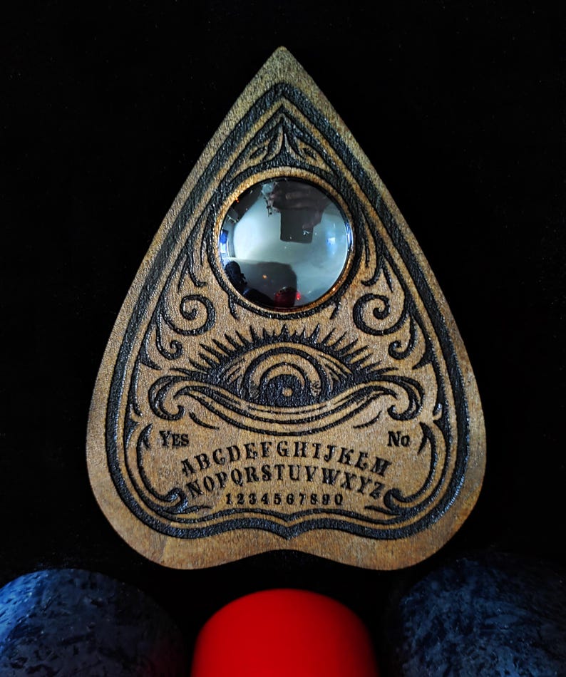 May include: A wooden Ouija board with engraved letters, numbers, and decorative elements. The board features a planchette and the words "Yes" and "No". The board is brown with black engraved details.