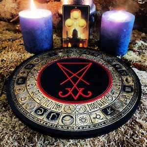 Puede incluir: Un tablero circular de madera negro y dorado con un símbolo de pentagrama rojo en el centro. El tablero está rodeado por un círculo de letras y símbolos dorados. Dos velas negras están a cada lado del tablero, y una carta de tarot está detrás del tablero.