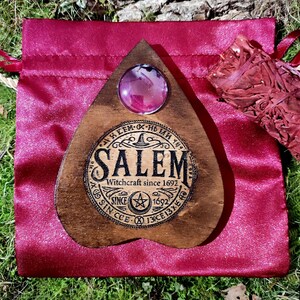 May include: A wooden planchette with a large, round, pink crystal and the word "Salem" in a circular design. The planchette is on a red satin bag with a bundle of dried herbs.