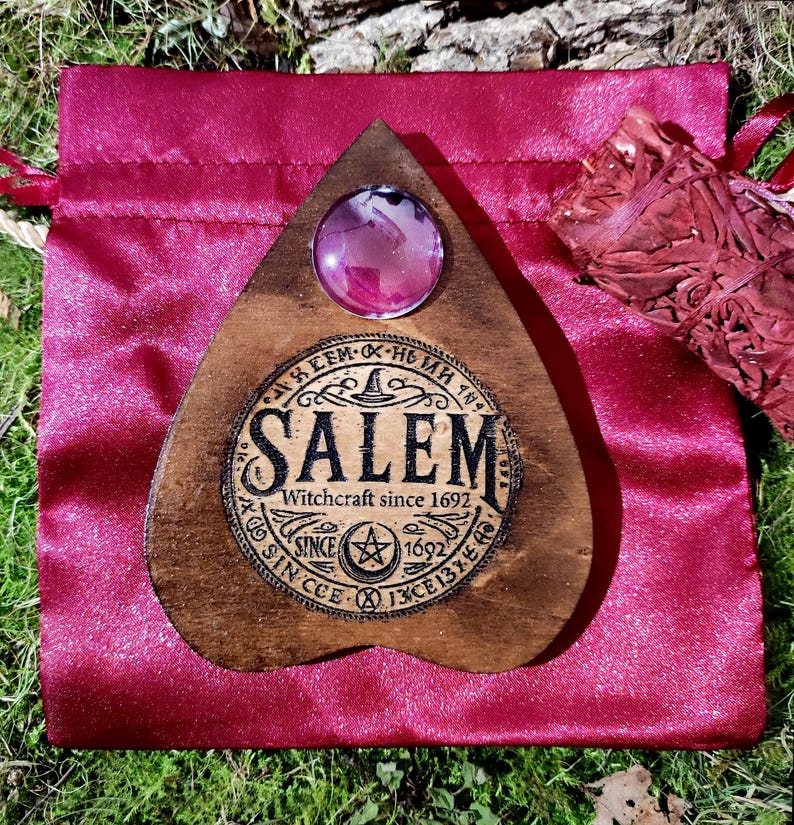 May include: A wooden planchette with a large, round, pink crystal and the word "Salem" in a circular design. The planchette is on a red satin bag with a bundle of dried herbs.