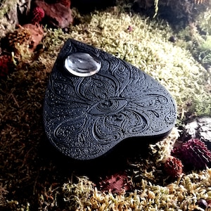Puede incluir: Tablero de madera negro en forma de corazón con un intrincado diseño de ojo y un cristal de vidrio transparente en la parte superior. El tablero está rodeado de musgo verde.
