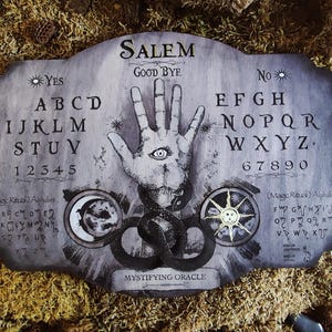 K&ouml;nnte beinhalten: Ein Ouija-Brett aus Holz mit einem schwarz-wei&szlig;en Design. Das Brett zeigt eine Hand mit einem Auge, einen zunehmenden Mond und eine Sonne. Das Brett hat die Buchstaben des Alphabets, Zahlen und die W&ouml;rter "Ja", "Nein" und "Leb wohl". Der Text "Salem Mystifying Oracle" ist auf dem Brett gedruckt.