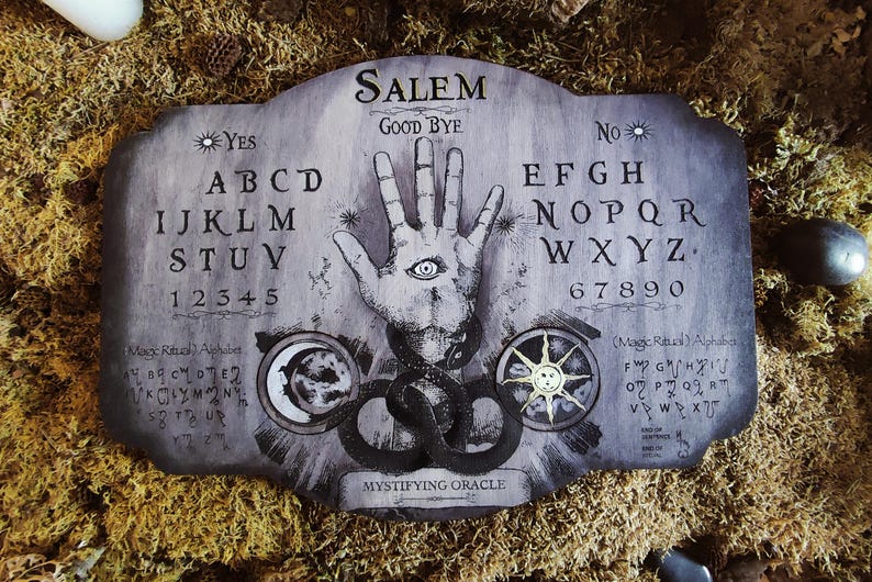 K&ouml;nnte beinhalten: Ein Ouija-Brett aus Holz mit einem schwarz-wei&szlig;en Design. Das Brett zeigt eine Hand mit einem Auge, einen zunehmenden Mond und eine Sonne. Das Brett hat die Buchstaben des Alphabets, Zahlen und die W&ouml;rter "Ja", "Nein" und "Leb wohl". Der Text "Salem Mystifying Oracle" ist auf dem Brett gedruckt.