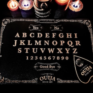 May include: A black Ouija board with gold lettering, featuring the alphabet, numbers, and the words "Yes," "No," and "Good Bye." The planchette is also black with the word "Ouija" and "Since 1891." Lit candles are in the background.
