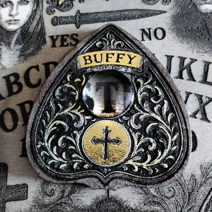 May include: A decorative planchette with the word "BUFFY" in gold lettering. The planchette is black with gold accents and a cross design. The letter "T" is visible through a clear lens.