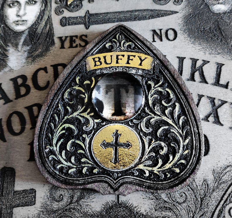 May include: A decorative planchette with the word "BUFFY" in gold lettering. The planchette is black with gold accents and a cross design. The letter "T" is visible through a clear lens.