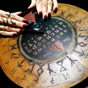 May include: A circular wooden Ouija board with a black and gold design, featuring letters, numbers, and symbols. A planchette rests on the board, with hands and long, dark red and black nails. The board has a vintage, mystical aesthetic.