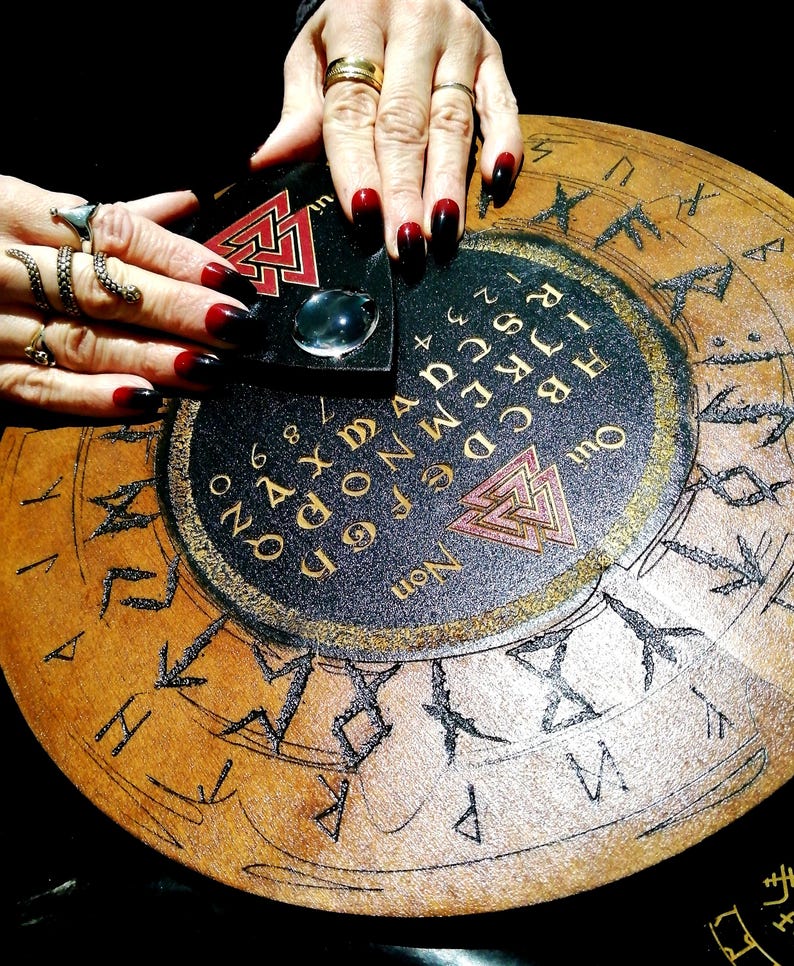 May include: A circular wooden Ouija board with a black and gold design, featuring letters, numbers, and symbols. A planchette rests on the board, with hands and long, dark red and black nails. The board has a vintage, mystical aesthetic.