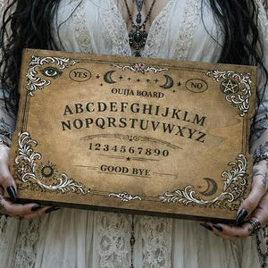 Op de afbeelding: Een rechthoekig houten Ouija-bord met een verweerde beige afwerking, voorzien van zwarte letters van het alfabet, de cijfers nul tot en met negen, de woorden YES, NO en GOOD BYE, en decoratieve hoekversieringen met hemelse symbolen.