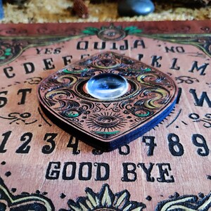May include: A wooden Ouija board with a planchette featuring a decorative eye design. The board has a dark brown background with gold and green accents. The letters, numbers, and words "YES", "NO", "GOOD BYE", and "OUIJA" are printed in black.