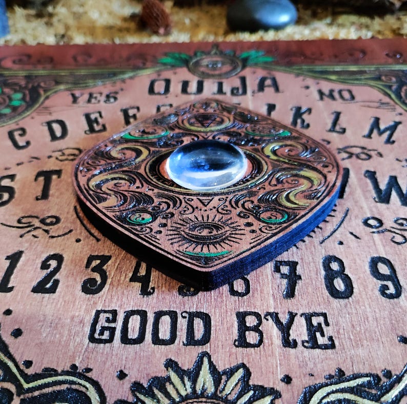 May include: A wooden Ouija board with a planchette featuring a decorative eye design. The board has a dark brown background with gold and green accents. The letters, numbers, and words "YES", "NO", "GOOD BYE", and "OUIJA" are printed in black.