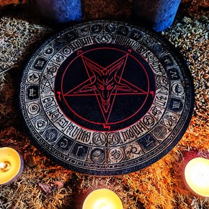 May include: A black and red wooden round coaster with a pentagram and a goat's head in the center. The pentagram is outlined in red and the goat's head is white. The coaster is surrounded by a circle of symbols and numbers.