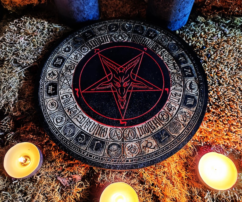 May include: A black and red wooden round coaster with a pentagram and a goat's head in the center. The pentagram is outlined in red and the goat's head is white. The coaster is surrounded by a circle of symbols and numbers.