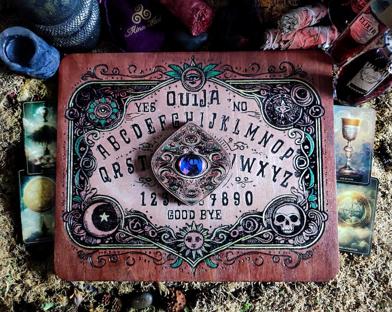 May include: A wooden Ouija board with a planchette featuring a blue gemstone. The board has a dark wood background with intricate green and gold designs. The letters of the alphabet, numbers 1-9 and 0, and the words "YES", "NO", and "GOOD BYE" are printed on the board.