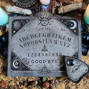 May include: A gray Ouija board with the text "OUIJA SINCE 1891" and the alphabet, numbers, and the words "YES", "NO", and "GOOD BYE". The board is surrounded by candles, cards, and sage bundles.