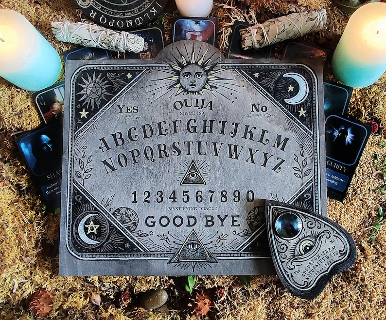 May include: A gray Ouija board with the text "OUIJA SINCE 1891" and the alphabet, numbers, and the words "YES", "NO", and "GOOD BYE". The board is surrounded by candles, cards, and sage bundles.