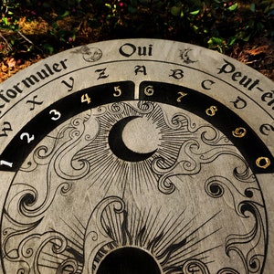 May include: A round, wooden divination board with etched celestial and floral designs. The board features the words "Oui" and "Peut-&ecirc;tre", along with numbers and letters. A crescent moon and sun are also etched into the board.