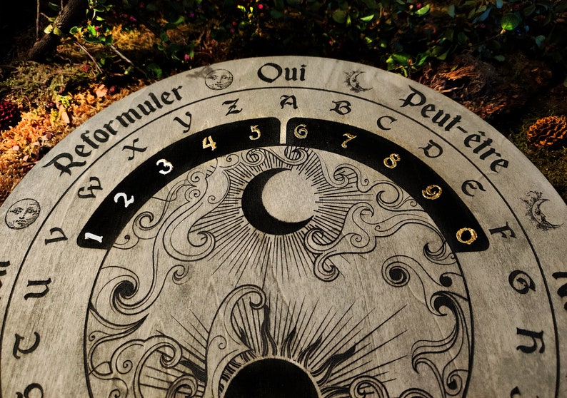 May include: A round, wooden divination board with etched celestial and floral designs. The board features the words "Oui" and "Peut-&ecirc;tre", along with numbers and letters. A crescent moon and sun are also etched into the board.