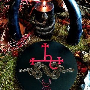 May include: A black circular altar cloth with a red inverted pentagram and a gold snake wrapped around it. The snake's tail is curled into a heart shape. Two red crescent moons are below the snake. The altar cloth is surrounded by moss, leaves, and a lit candle.