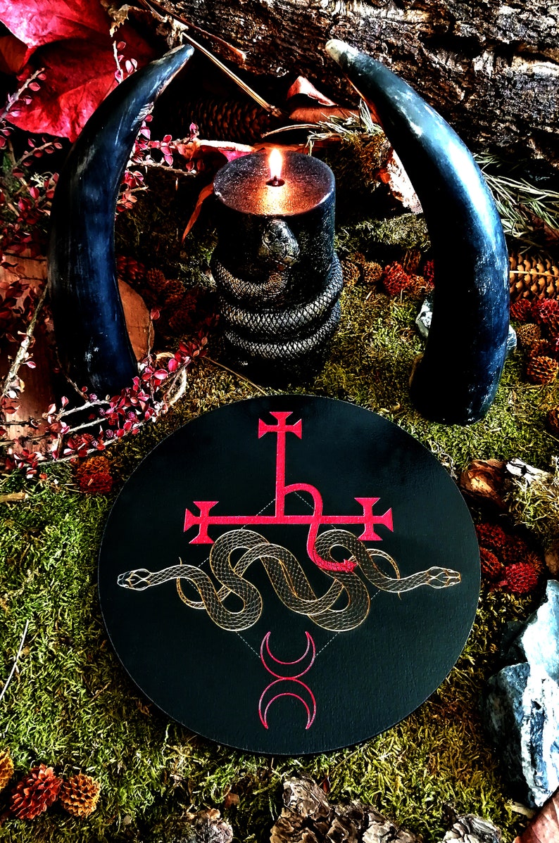 May include: A black circular altar cloth with a red inverted pentagram and a gold snake wrapped around it. The snake's tail is curled into a heart shape. Two red crescent moons are below the snake. The altar cloth is surrounded by moss, leaves, and a lit candle.