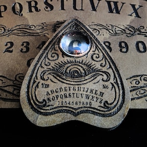 May include: A wooden planchette for a Ouija board, featuring a black and white design of an eye with a decorative border. The planchette has a clear glass orb in the center with the number "6" visible.