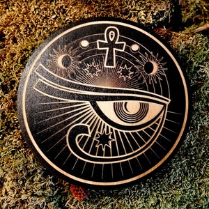 May include: Black wooden round coaster with a white design of an eye with an ankh symbol above it and stars and moons around it.