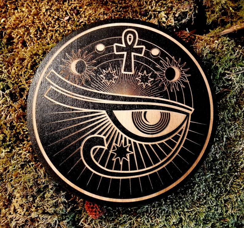 May include: Black wooden round coaster with a white design of an eye with an ankh symbol above it and stars and moons around it.
