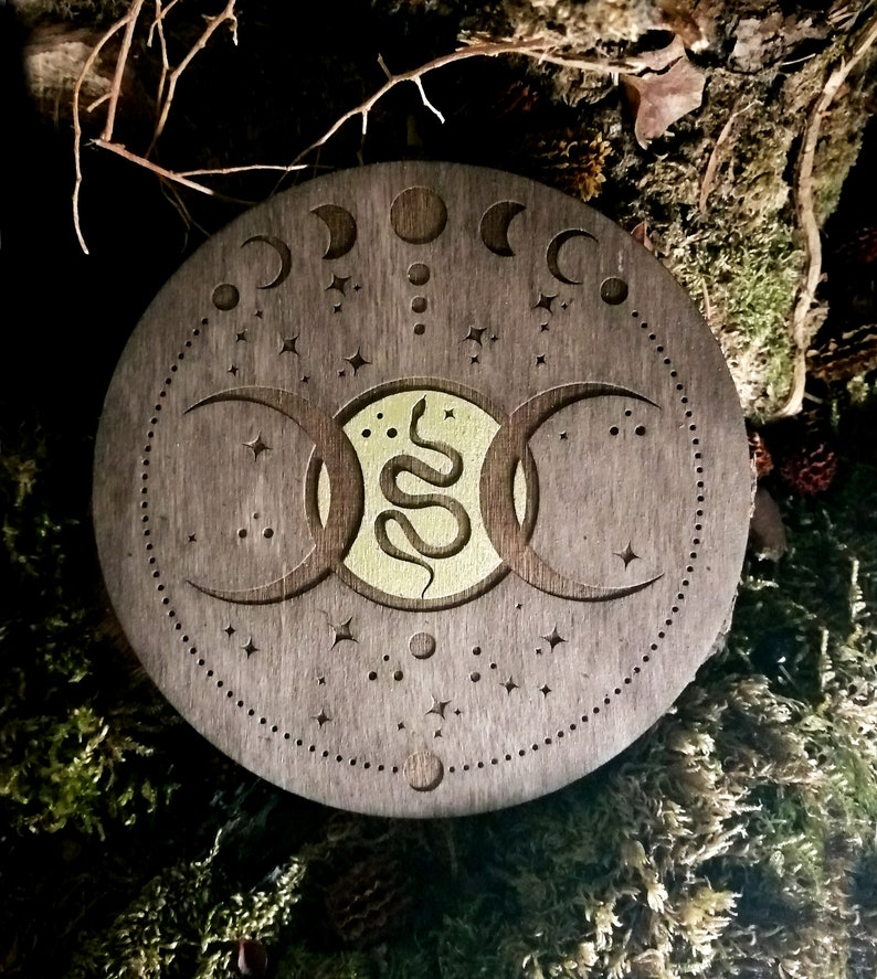 May include: A wooden circle with a triple moon design and a snake in the center. The circle is decorated with stars and moons.
