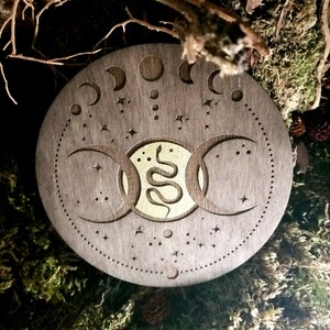 May include: A round wooden coaster with a triple moon symbol and a snake design in gold. The coaster is decorated with stars and moons.
