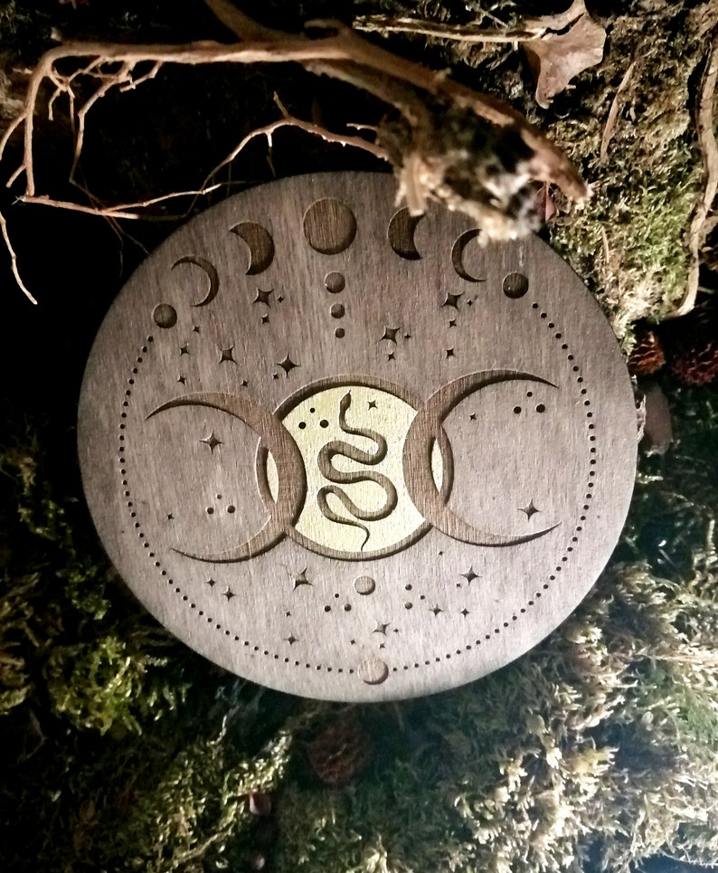 May include: A round wooden coaster with a triple moon symbol and a snake design in gold. The coaster is decorated with stars and moons.