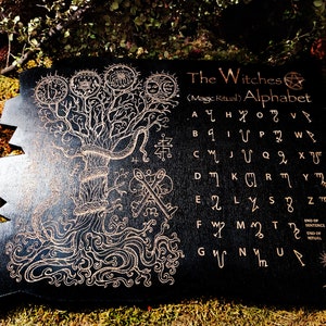 May include: A black wooden board with a gold-colored tree and a witch's alphabet. The alphabet is written in a stylized font and includes symbols for each letter. The text reads "The Witches (Magic Ritual) Alphabet".