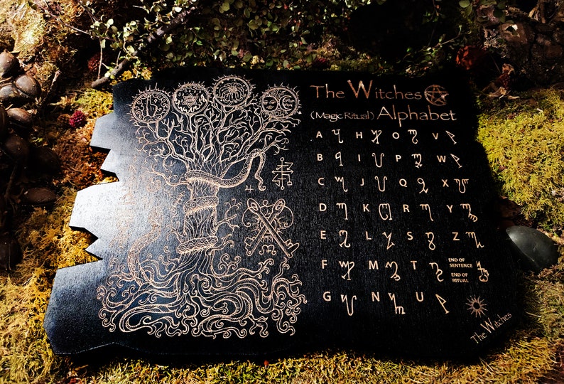 May include: A black wooden board with a gold-colored tree and a witch's alphabet. The alphabet is written in a stylized font and includes symbols for each letter. The text reads "The Witches (Magic Ritual) Alphabet".