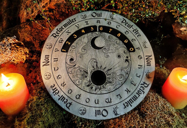 May include: A round, wooden divination board with etched letters, numbers, and symbols. The board features the words "Oui", "Non", and "Peut-&ecirc;tre", along with the numbers 1-9. The board is surrounded by candles and moss.