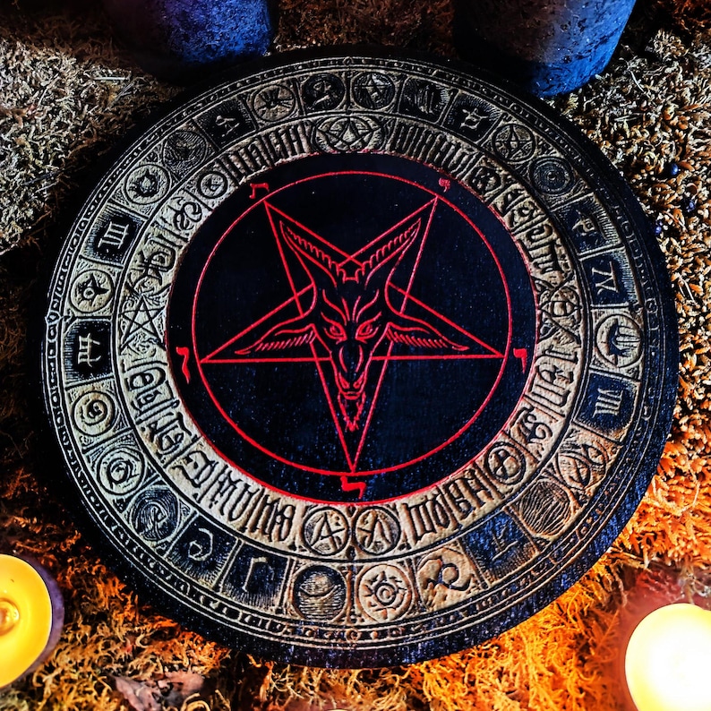 May include: A black wooden round coaster with a red pentagram and a goat's head in the center. The pentagram is surrounded by a circle of symbols and text.