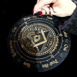 May include: A black and gold circular pendulum board with the words "Oui", "Peut-&ecirc;tre", "Non", "Reformuler", and "Divinum" in French and English. The board features a gold all-seeing eye design with a pendulum hanging above it.