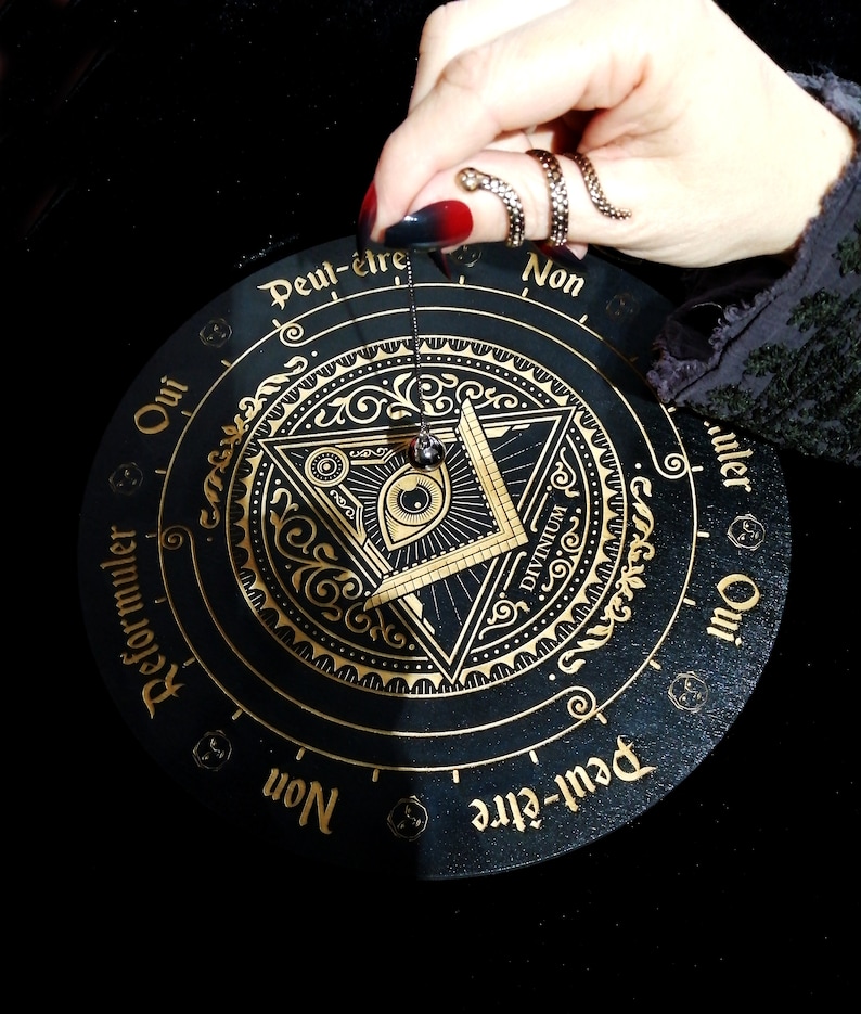 May include: A black and gold circular pendulum board with the words "Oui", "Peut-&ecirc;tre", "Non", "Reformuler", and "Divinum" in French and English. The board features a gold all-seeing eye design with a pendulum hanging above it.