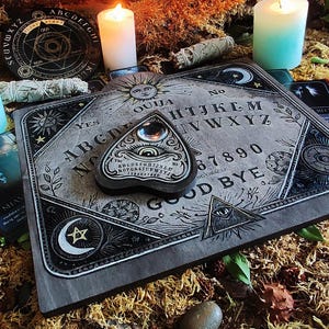 May include: A gray Ouija board with a planchette, surrounded by candles, tarot cards, and dried moss. The board features the alphabet, numbers, and the words "YES" and "GOOD BYE". The planchette has an eye design.