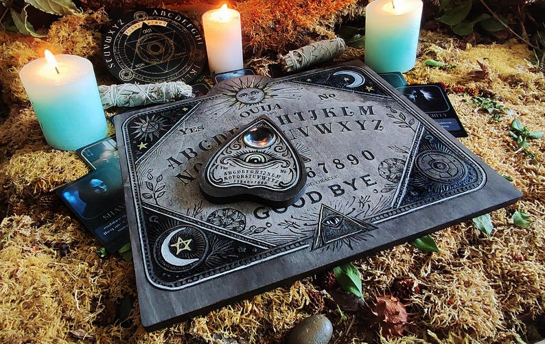 May include: A gray Ouija board with a planchette, surrounded by candles, tarot cards, and dried moss. The board features the alphabet, numbers, and the words "YES" and "GOOD BYE". The planchette has an eye design.