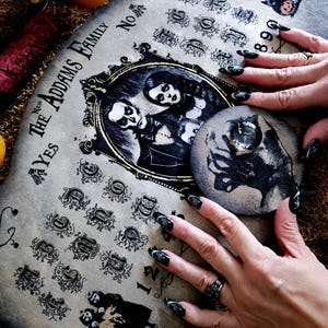 May include: A vintage-style Ouija board featuring the text "The Addams Family" and a portrait of the family. The board displays letters, numbers, and symbols. A planchette with a cat illustration is on the board. The board is made of wood with a distressed finish.