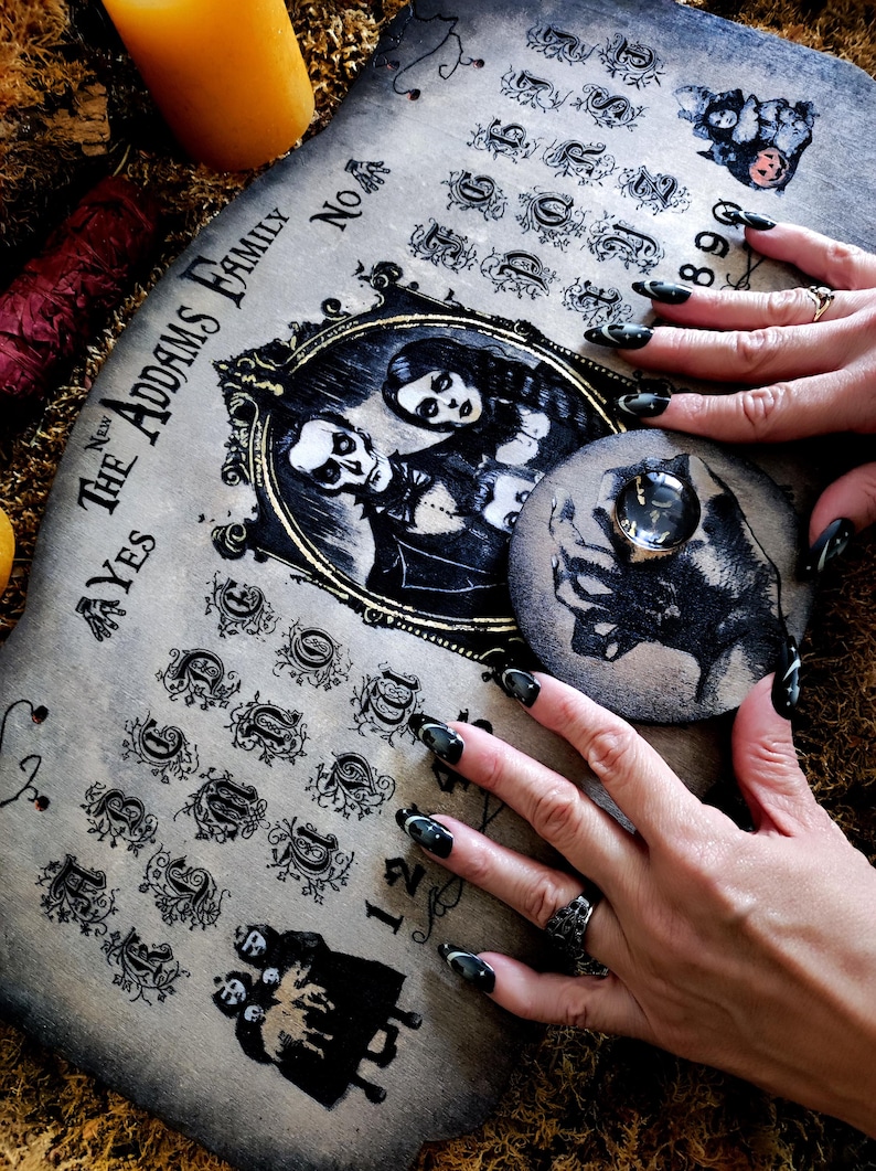 May include: A vintage-style Ouija board featuring the text "The Addams Family" and a portrait of the family. The board displays letters, numbers, and symbols. A planchette with a cat illustration is on the board. The board is made of wood with a distressed finish.