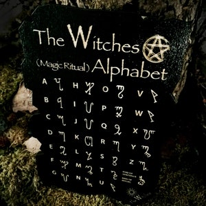 May include: Black wooden sign with white text that reads "The Witches (Magic Ritual) Alphabet". The sign features a pentagram symbol and a white alphabet with corresponding symbols.